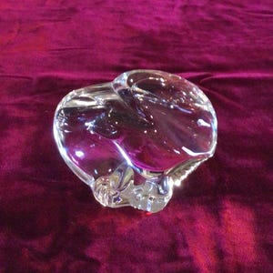 May include: A clear glass ashtray with a heart-shaped design. The ashtray is made of thick, transparent glass and has a smooth, polished surface. The ashtray is sitting on a deep red velvet surface.