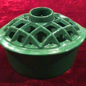 May include: A glossy, forest green ceramic pot with a matching lid. The lid features a raised, open-lattice diamond pattern and a central circular knob. The pot has two small, curved side handles and a smooth, solid-colored base body.
