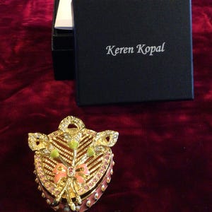 May include: A heart-shaped trinket box with a gold-tone finish, adorned with pink and clear crystals. The box features a pink enamel base and a decorative bow. The black box with the text "Keren Kopal" is in the background.