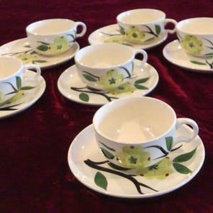May include: A set of six white ceramic teacups and saucers. Each cup and saucer features a floral design with yellow flowers and green leaves on black branches. The cups have a curved handle.