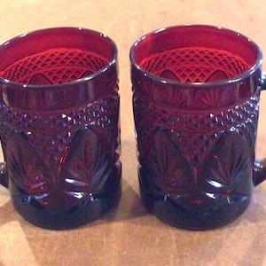 May include: Two ruby red glass mugs with a textured, diamond-patterned design. Each mug has a sturdy handle and a detailed pattern on the body. The mugs are made of a dark red glass, perfect for serving hot beverages.