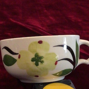 May include: White ceramic teacup with a large floral design in shades of green and yellow, accented with black and red details. The cup has a curved handle and a wide, shallow bowl, perfect for serving tea or soup.
