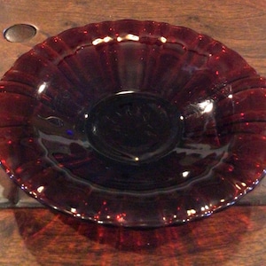 May include: A ruby red glass bowl with a scalloped rim and two small handles. The bowl has a circular shape with a slightly raised center. The glass has a glossy finish, reflecting light and creating highlights.