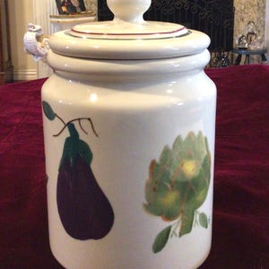 May include: A white ceramic jar with a lid, featuring hand-painted illustrations of an eggplant and an artichoke. The jar has a handle shaped like a bird and is decorated with red and green stripes around the lid.
