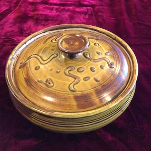 May include: A round, lidded ceramic dish with a brown and yellow glaze. The lid features a small knob and is decorated with brown, wavy lines and dots. The dish has a striped pattern around the base.