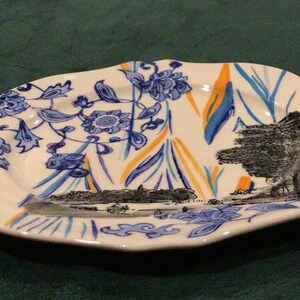 May include: A white oval ceramic serving dish with a blue and orange floral design. The dish features a black and white landscape scene with trees and a river.