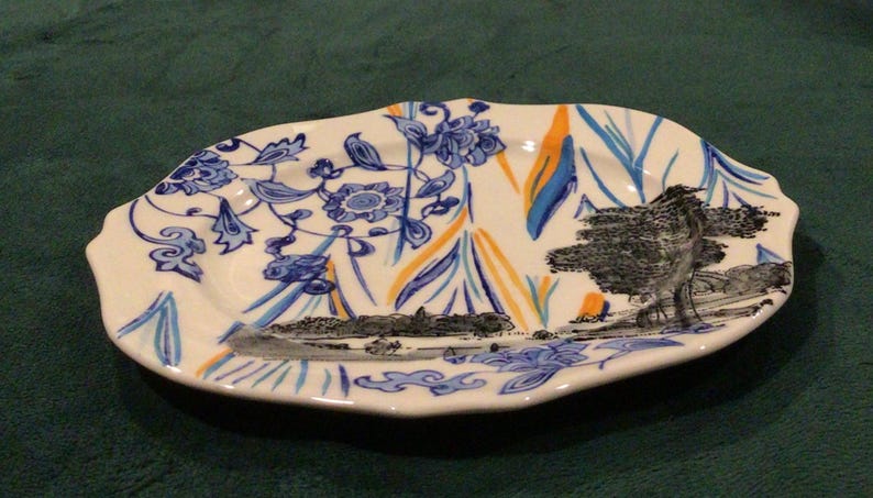 May include: A white oval ceramic serving dish with a blue and orange floral design. The dish features a black and white landscape scene with trees and a river.