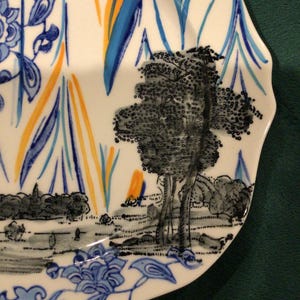 May include: A white ceramic plate with a blue and yellow floral design. The plate features a black and white illustration of a tree and a landscape.