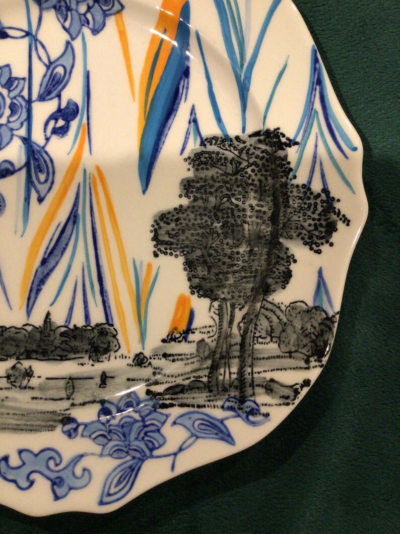 May include: A white ceramic plate with a blue and yellow floral design. The plate features a black and white illustration of a tree and a landscape.