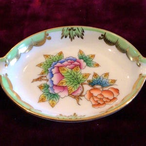 May include: An oval porcelain dish featuring a central floral motif with pink, blue, and orange flowers surrounded by green leaves. The rim is decorated with a light green border and gold accents, and the dish includes two small side notches.