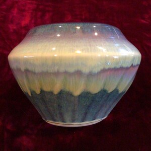 May include: A ceramic vase with a wide, rounded body and a narrow opening. The surface features a reactive glaze with a color gradient transitioning from a deep teal at the base to a creamy off-white in the middle and a soft lavender at the rim.