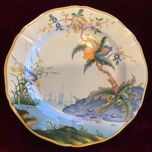 May include: A white ceramic plate with a scalloped edge and a yellow border. The plate features a hand-painted design of a bird, a tree with fruit, and a landscape with water and blue accents. The plate is approximately 12 inches in diameter.