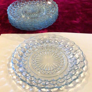 May include: A set of light blue glass plates featuring a raised hobnail pattern along the rim and a central sunburst design. The plates have a scalloped edge and a translucent finish, with one plate displayed in the foreground and a stack of four behind.