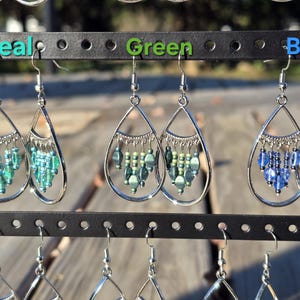 May include: Teal, green, and blue beaded earrings displayed on a black earring stand. Each pair features a teardrop-shaped silver frame with dangling beads. The earrings are arranged in rows, showcasing their different colors.