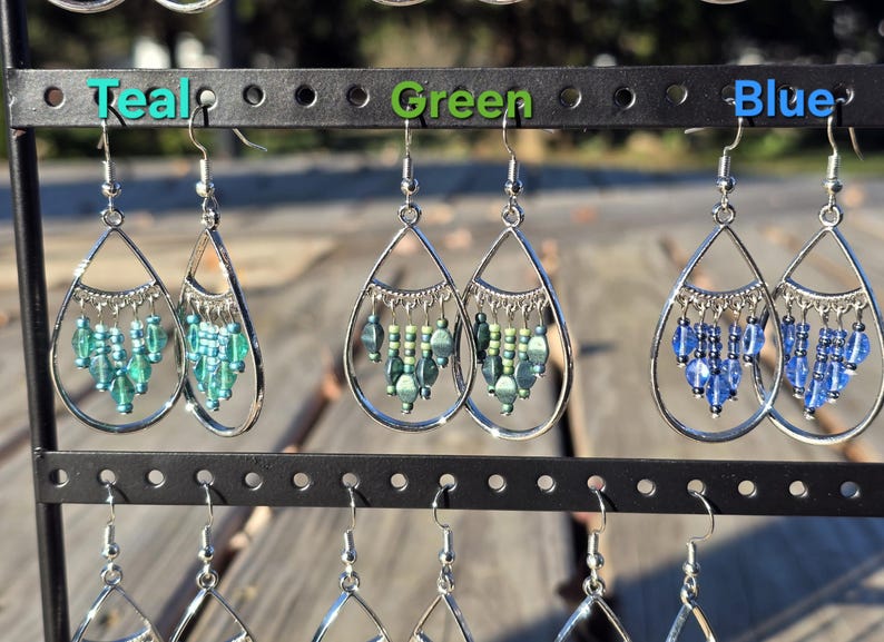 May include: Teal, green, and blue beaded earrings displayed on a black earring stand. Each pair features a teardrop-shaped silver frame with dangling beads. The earrings are arranged in rows, showcasing their different colors.
