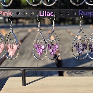 May include: Earrings in pink, lilac, and purple hang from a black display rack. Each earring features a silver teardrop frame with dangling beads. The word "Pink", "Lilac", and "Purple" are above each pair of earrings.