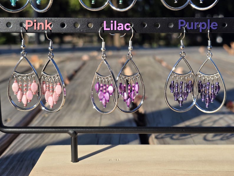 May include: Earrings in pink, lilac, and purple hang from a black display rack. Each earring features a silver teardrop frame with dangling beads. The word "Pink", "Lilac", and "Purple" are above each pair of earrings.