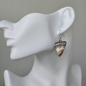 Puede incluir: Pendientes triangulares con dise&ntilde;o de piedra natural. Los pendientes presentan una mezcla de tonos marr&oacute;n, blanco y gris, con un gancho plateado. Se muestran en una cabeza de maniqu&iacute; blanca.