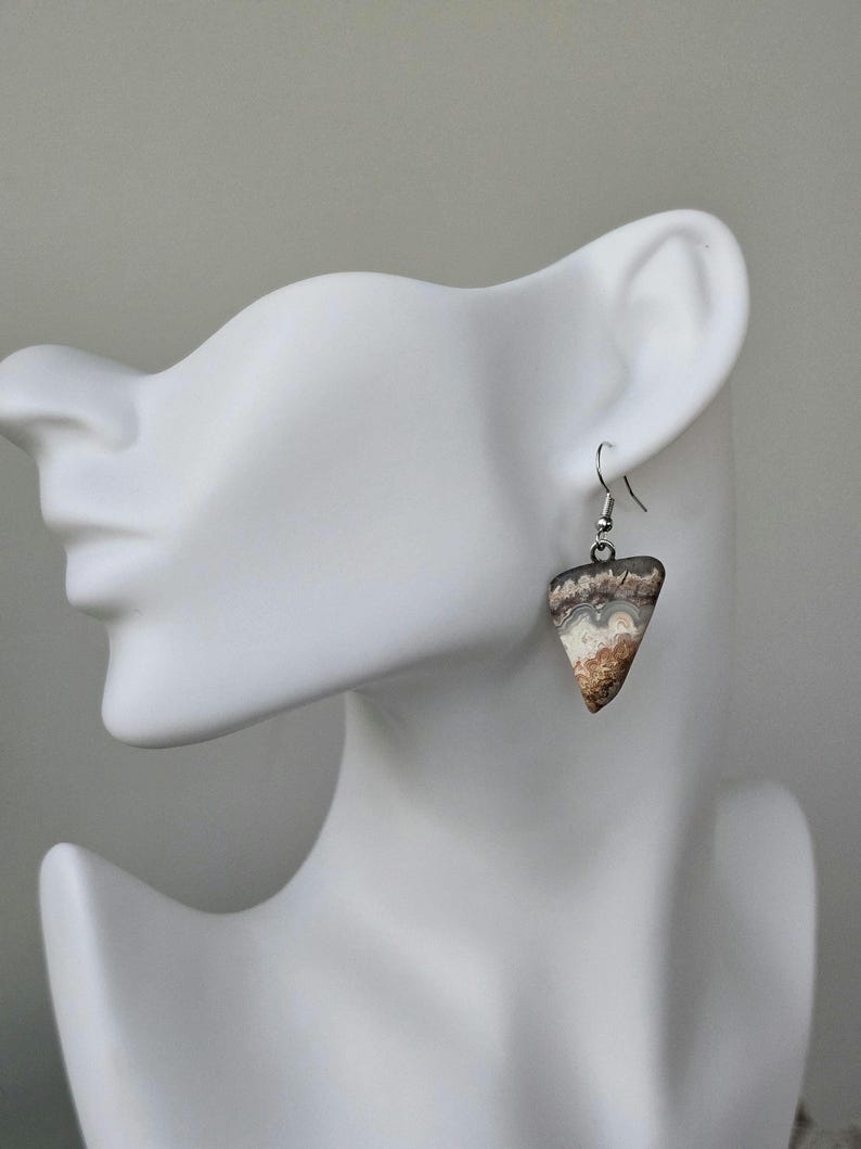 Puede incluir: Pendientes triangulares con dise&ntilde;o de piedra natural. Los pendientes presentan una mezcla de tonos marr&oacute;n, blanco y gris, con un gancho plateado. Se muestran en una cabeza de maniqu&iacute; blanca.