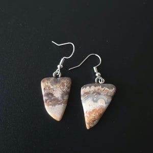 Puede incluir: Un par de pendientes artesanales con piedras triangulares pulidas que muestran patrones naturales en tonos beige, marr&oacute;n y gris. Los pendientes tienen ganchos para las orejas plateados, perfectos para el uso diario.