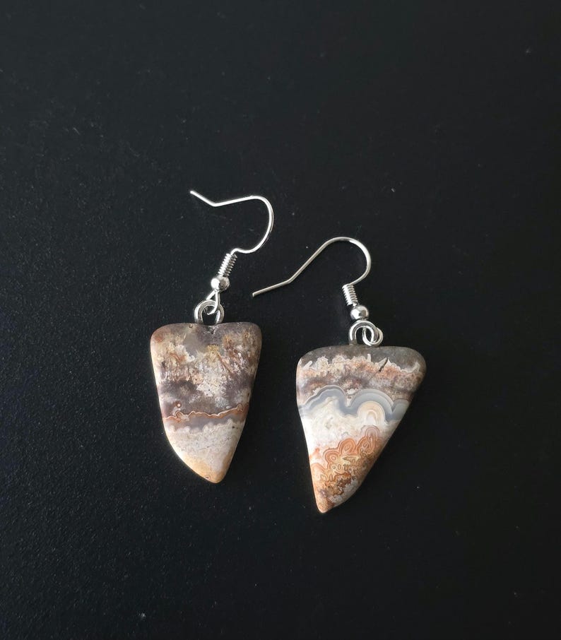 Puede incluir: Un par de pendientes artesanales con piedras triangulares pulidas que muestran patrones naturales en tonos beige, marr&oacute;n y gris. Los pendientes tienen ganchos para las orejas plateados, perfectos para el uso diario.