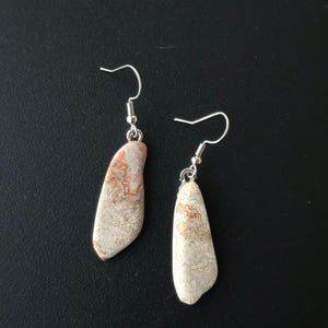 Puede incluir: Un par de pendientes colgantes con colgantes de piedra natural. Los pendientes tienen ganchos plateados y peque&ntilde;as gotas de piedra estampada en tonos beige, crema y marr&oacute;n rojizo. Los pendientes se muestran sobre un fondo negro.