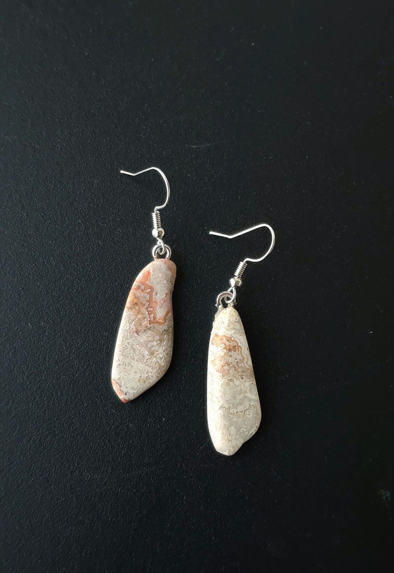 Puede incluir: Un par de pendientes colgantes con colgantes de piedra natural. Los pendientes tienen ganchos plateados y peque&ntilde;as gotas de piedra estampada en tonos beige, crema y marr&oacute;n rojizo. Los pendientes se muestran sobre un fondo negro.