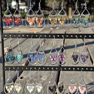 Op de afbeelding: Een display van verschillende druppelvormige oorbellen met kralen. Elke oorbel heeft een zilverkleurig frame en een waterval van kleurrijke kralen in tinten rood, oranje, goud, groen, blauw, paars, zwart, wit en roze. De oorbellen hangen aan een zwart metalen rek.