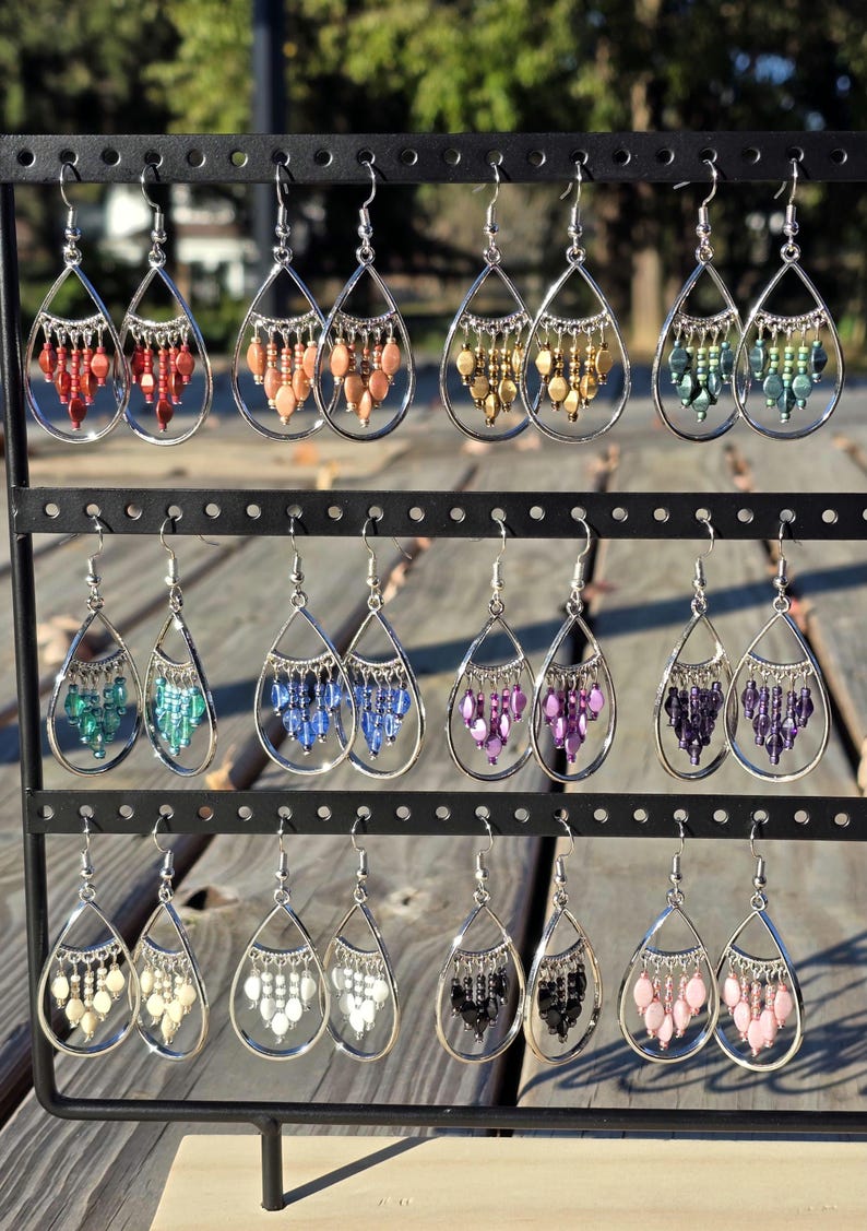 May include: A display of various beaded teardrop earrings. Each earring features a silver-tone frame with a cascade of colorful beads in shades of red, orange, gold, green, blue, purple, black, white, and pink. The earrings are hanging on a black metal rack.