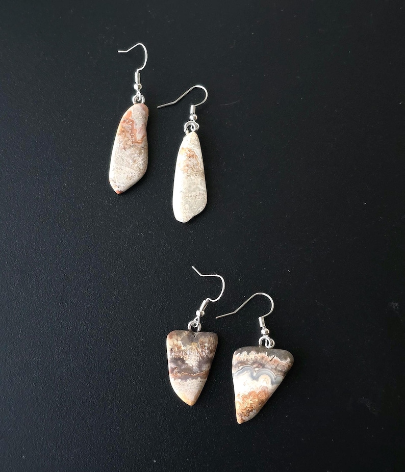 Puede incluir: Cuatro pares de pendientes de piedra con ganchos plateados. Los pendientes tienen una forma natural y org&aacute;nica y presentan una mezcla de tonos beige, marr&oacute;n y blanco. Los pendientes se muestran sobre una superficie negra.
