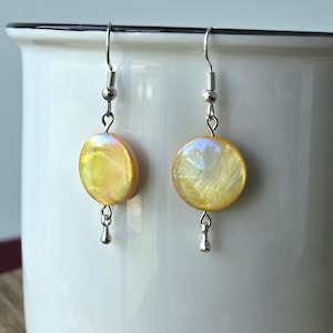 May include: A pair of dangle earrings with iridescent, yellow-gold, oval-shaped pendants. The earrings have silver-colored hooks and small, teardrop-shaped charms hanging below the pendants. The earrings are displayed against a white background.