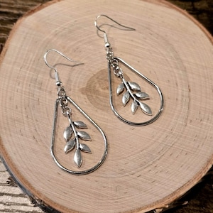 May include: Silver-toned drop earrings featuring a teardrop-shaped frame with a dangling leaf design. The earrings have a hook closure and are displayed on a wooden surface. The earrings are a nature-inspired jewelry item.