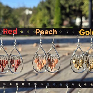 May include: Three pairs of teardrop earrings in red, peach, and gold. Each pair has a silver-tone frame with dangling beads. The earrings are displayed on a black earring stand with the colors labeled above.