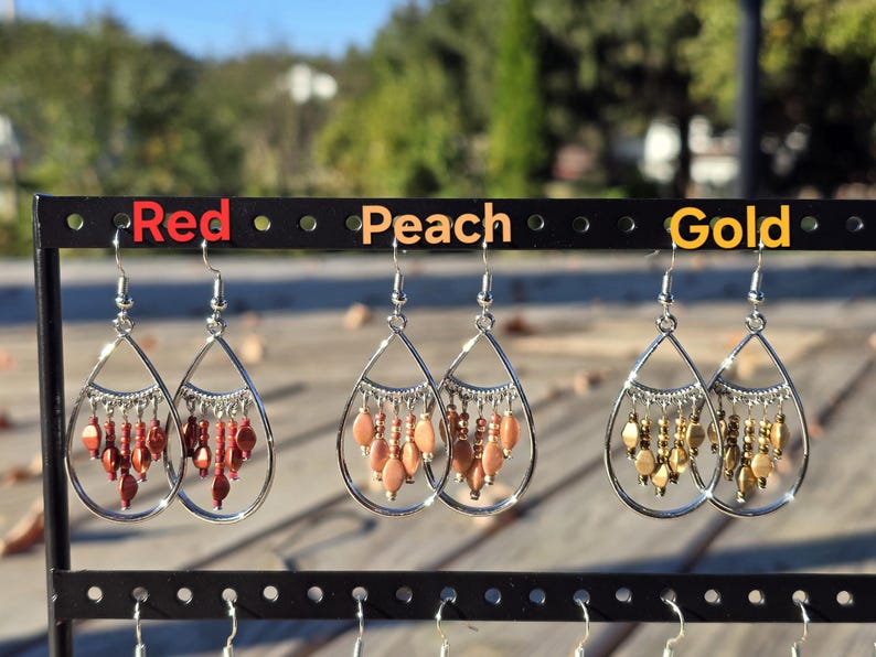 May include: Three pairs of teardrop earrings in red, peach, and gold. Each pair has a silver-tone frame with dangling beads. The earrings are displayed on a black earring stand with the colors labeled above.