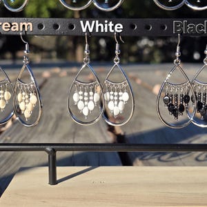 May include: A display of three pairs of teardrop earrings in cream, white, and black. Each earring features a silver-tone frame with dangling beads. The earrings are suspended from a black metal rack, with the colors labeled above.