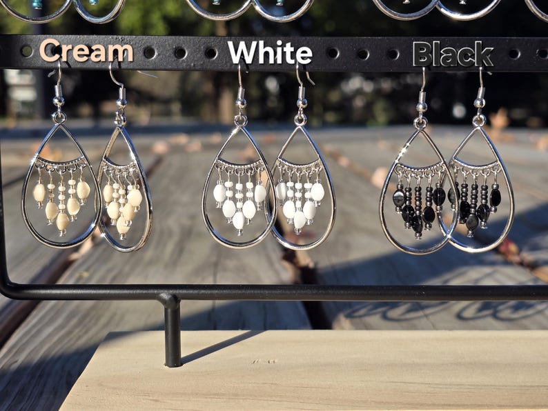 May include: A display of three pairs of teardrop earrings in cream, white, and black. Each earring features a silver-tone frame with dangling beads. The earrings are suspended from a black metal rack, with the colors labeled above.