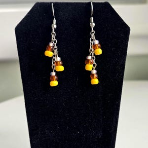 May include: Dangle earrings featuring a candy corn design. Each earring has a silver-tone hook and chain, with clusters of yellow, orange, and white beads. The earrings are displayed on a black velvet stand.