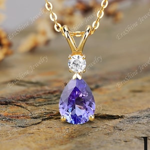 Pode incluir: Um colar de ouro com um pingente de pedra preciosa roxa escura em forma de pêra. O pingente é acentuado com uma pequena pedra preciosa redonda e transparente acima. O colar é exibido em uma superfície marrom texturizada.