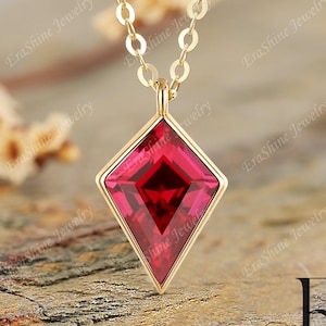 May include: A gold necklace featuring a kite-shaped pendant with a deep red gemstone set in a thin gold frame. The pendant hangs from a delicate gold chain. The text EraShine Jewelry and the letters ES are visible in the bottom right corner.