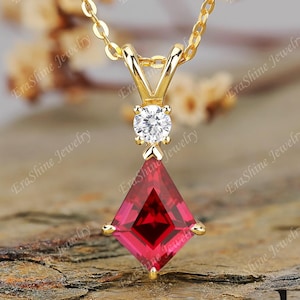 May include: A gold-colored necklace with a pendant featuring a vibrant red, square-cut gemstone. Above the red stone is a small, round, clear gemstone. The necklace is displayed on a textured, brown surface.