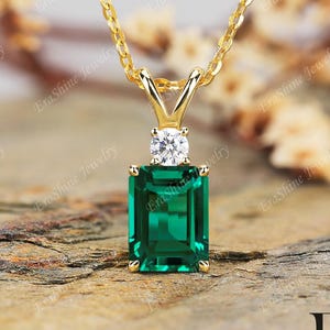 May include: A gold necklace with a rectangular emerald-cut green gemstone pendant. A small, round, clear gemstone sits above the green stone. The necklace is on a gold chain.