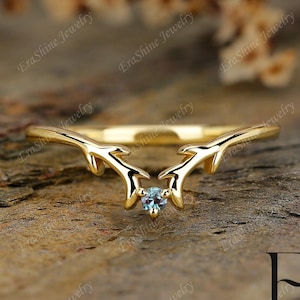 May include: A gold ring featuring a delicate antler design with a small blue gemstone at its centre. The ring is displayed against a wooden backdrop.
