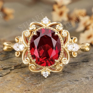 Puede incluir: Un anillo de oro con una gran piedra preciosa de color rojo intenso y corte ovalado en el centro, rodeada por un marco de filigrana ornamentado. El engaste incluye cuatro pequeñas piedras de acento redondas y claras en los extremos.