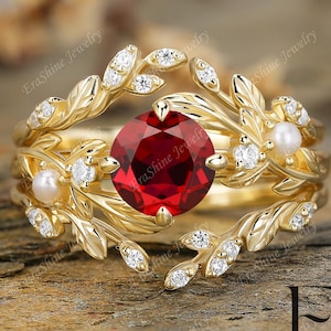 May include: A gold-colored ring with a large, round, red gemstone at the center. The ring features a floral design with small white gemstones and pearls. The band is adorned with leaf-like details. The ring is set against a blurred background.