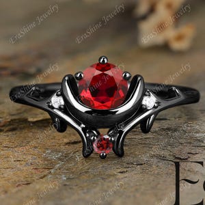 May include: A black ring with a large, round, red gemstone at the center, flanked by smaller red and white gemstones. The ring has an ornate, curved design with the text "EraShine Jewelry".