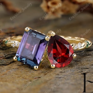 Könnte beinhalten: Ein goldener Ring mit zwei Edelsteinen: einem rechteckigen lila-blauen Stein und einem birnenförmigen roten Stein. Der Ring hat ein strukturiertes Band mit kleinen Diamanten und grünen Akzenten. Der Markenname "EraShine Jewelry" ist sichtbar.