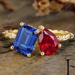 Op de afbeelding: Een gouden ring met een grote, smaragdgeslepen blauwe edelsteen en een peervormige rode edelsteen. De band heeft een textuurontwerp met kleine diamanten en groene accenten. De ring ligt op een houten oppervlak.