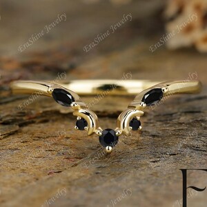 May include: A delicate gold wedding band with a V-shaped design and five black gemstones. The band is set against a wooden background.