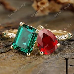 Puede incluir: Un anillo de oro con una esmeralda verde de talla esmeralda y una piedra roja en forma de pera. El anillo tiene una banda retorcida con pequeñas piedras de acento y el texto "EraShine Jewelry".