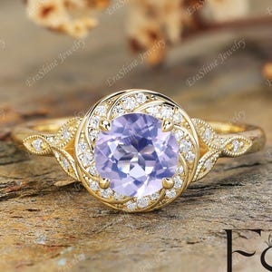 Può includere: Un anello d'oro con una gemma rotonda, viola chiaro, circondata da piccole pietre trasparenti. L'anello presenta dettagli intricati sulla fascia e intorno alla pietra centrale. È posto su una superficie marrone strutturata.